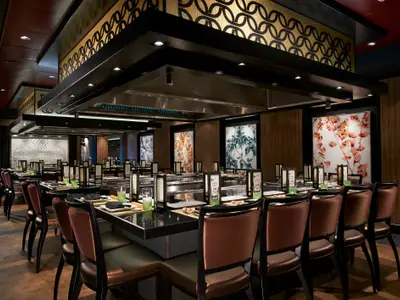Ein weitläufiger Blick in das elegante Teppanyaki-Restaurant an Bord der Norwegian Escape, mit mehreren dunklen Tischen und braunen Stühlen. Die Tische sind gedeckt mit Tellern, Gläsern und Menükarten. Über den Tischen hängt eine massive, schwarz-goldene Deckenkonstruktion mit integrierten Lichtern und Dunstabzügen. Die Wände sind mit Holzpaneelen und beleuchteten Kunstwerken mit Blattmotiven verziert. Es sind keine Personen zu sehen.