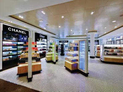 Heller, moderner Duty-Free Shop an Bord der Norwegian Escape. Beleuchtete Regale präsentieren Parfüm und Kosmetik von Marken wie Chanel und Dior. "Duty Free Exclusive"-Schild. Gemusterter Boden. Ohne Personen.