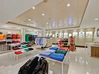 Ein gut beleuchteter Lacoste Shop an Bord der Norwegian Escape, der bunte Poloshirts, Hemden und Handtaschen auf Regalen und Tischen präsentiert.