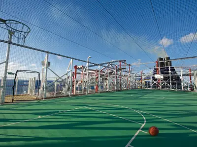 Ein weitläufiger Basketballplatz an Bord der Norwegian Escape, umgeben von Fangnetzen. Im Hintergrund sind Teile der Schiffsinfrastruktur, Schornsteine und das weite blaue Meer zu sehen. Der Himmel ist klar mit wenigen Wolken.