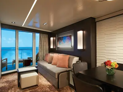 Innenansicht einer eleganten Suite auf der Norwegian Escape. Ein gemütlicher Wohnbereich mit Sofa, Couchtisch und Meerblick vom Balkon. Helle Tagesbeleuchtung.