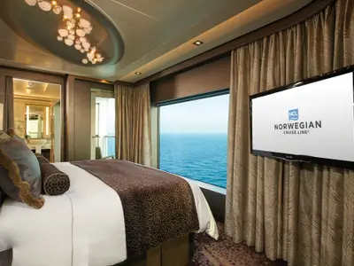 Eine elegante Suite an Bord der Norwegian Escape mit einem grossen Doppelbett, Meerblick durch ein Panoramafenster und einem Flachbildfernseher. Im Hintergrund ist ein Badezimmer zu sehen.