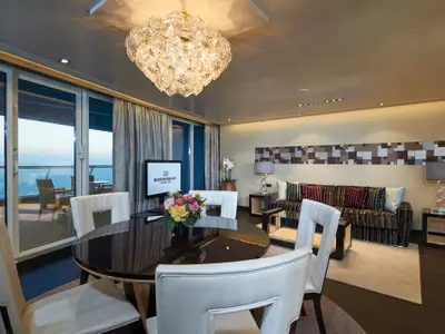Ein luxuriöses Wohnzimmer einer Suite an Bord der Norwegian Escape, mit elegantem Essbereich, Sofa, Couchtischen und großen Glastüren, die auf einen Balkon mit Meerblick führen. Ein opulent leuchtender Kronleuchter hängt von der Decke.
