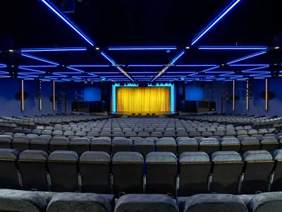 Blick in das leere, moderne Theater der Norwegian Escape. Blaue LED-Lichtleisten am Decke und blaue Wände. Die Bühne ist mit einem goldenen Vorhang geschmückt.