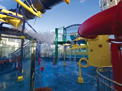 Ein lebhafter, farbenfroher AquaPark an Bord der Norwegian Escape, mit blauen Wasserspielen, gelben Rutschen und roten Spritzdüsen, unter strahlend blauem Himmel. Ohne Personen.