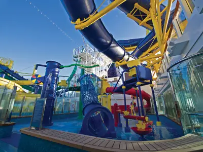 Der lebhafte Kids AquaPark an Bord der Norwegian Escape. Bunte Wasserrutschen, Eimer und Spritzdüsen füllen die blauen Becken. Leere Anlage bei Tag unter blauem Himmel.
