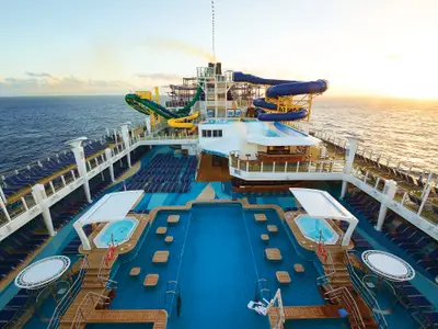 Panoramablick auf den AquaPark an Bord der Norwegian Escape bei Sonnenuntergang. Zu sehen sind mehrere Pools, Whirlpools, zahlreiche blaue Liegestühle und große, farbige Wasserrutschen, die sich über mehrere Decks erstrecken. Das Meer und der Horizont mit tiefstehender Sonne bilden den Hintergrund.