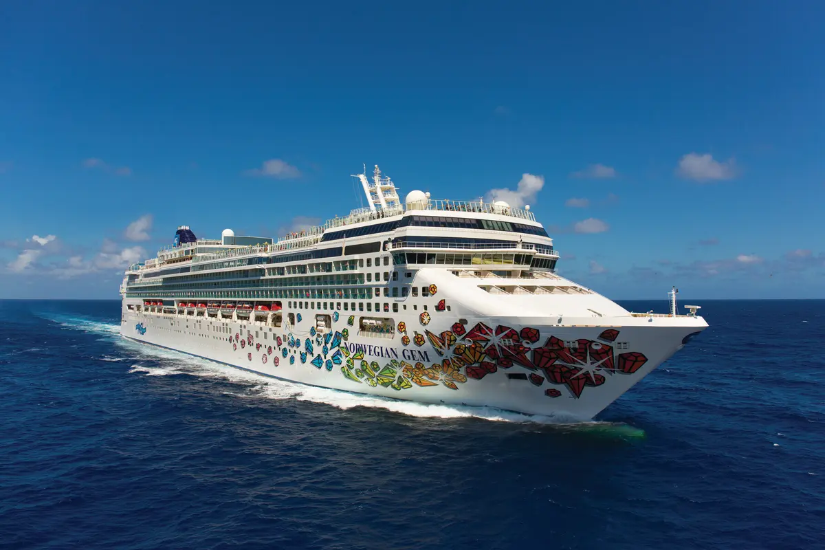 Luftbild der Norwegian Gem, die auf tiefblauem Meer fährt. Das weiße Kreuzfahrtschiff hat eine bunte Edelstein-Rumpfbemalung. Blauer Himmel mit Wolken.