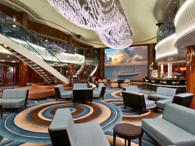 Das Atrium der Norwegian Gem mit grosser Freitreppe, blauen Sitzgruppen und Bar. Ein riesiger Bildschirm zeigt ein Kreuzfahrtschiff im Sonnenuntergang auf See.