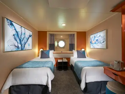 Die Aussenkabine an Bord der Norwegian Gem zeigt zwei Betten mit blauen Akzenten, ein Bullauge und moderne Kunst. Ein Schreibtisch mit Getränken ist rechts sichtbar.