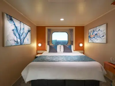 Moderne Aussenkabine an Bord der Norwegian Gem mit Doppelbett, blau gemusterter Tagesdecke und Bullaugenfenster. Zwei abstrakte, blaue Kunstwerke zieren die Wände.