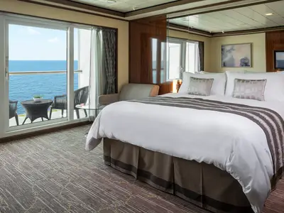 Blick in eine moderne Balkonkabine an Bord der Norwegian Gem. Ein grosses Doppelbett mit weisser Bettwäsche und grauer Decke füllt den Vordergrund. Im Hintergrund öffnet sich eine Schiebetür zu einem Balkon mit Meerblick, zwei Stühlen und einem Tisch. Tageslicht durchflutet den Raum.