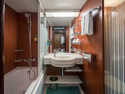 Badezimmer einer Balkonkabine an Bord der Norwegian Gem. Es zeigt eine Dusche über einer Badewanne, ein Waschbecken mit Spiegel und Handtücher. Die Wände sind aus dunklem Holz.