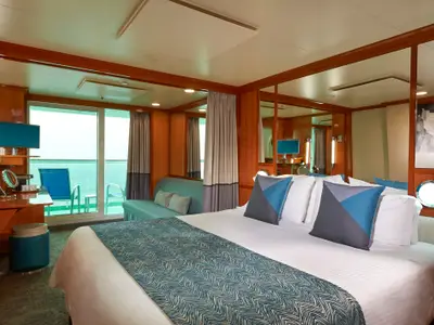 Eine geräumige, moderne Balkonkabine an Bord der Norwegian Gem mit einem großen Doppelbett, einer Sitzecke mit Sofa und einem Schreibtisch. Der Balkon mit zwei Stühlen ist durch eine Glastür zugänglich.
