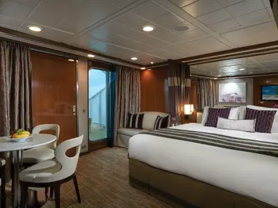 Geräumige Balkonkabine an Bord der Norwegian Gem mit großem Bett, Nachttischen, einem kleinen Esstisch mit Stühlen und Zugang zum Balkon mit Meerblick. Lila Akzente in Kissen und Vorhängen.