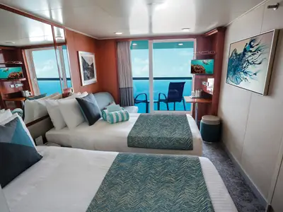 Blick in eine moderne Balkonkabine der Norwegian Gem. Zwei Einzelbetten mit blauen Akzenten, ein Sofa und ein Balkon mit Meerblick sind zu sehen.