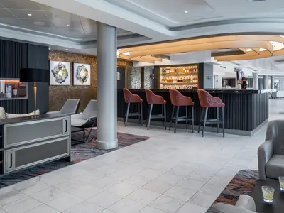 Eine moderne Bar-Lounge an Bord der Norwegian Gem, die Maltings Whiskey Bar. Sie zeigt eine lange Bar mit Barhockern in warmem Rotton, einem Regal voller Spirituosen, stilvolle Wandkunst und helle Marmorböden.