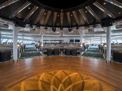 Die Spinnaker Lounge an Bord der Norwegian Gem. Eine breite Holzfläche mit Parkettmuster im Vordergrund, umgeben von geschwungenen Sitzgruppen und einer Bar im Hintergrund. Scheinwerfer an der Decke.