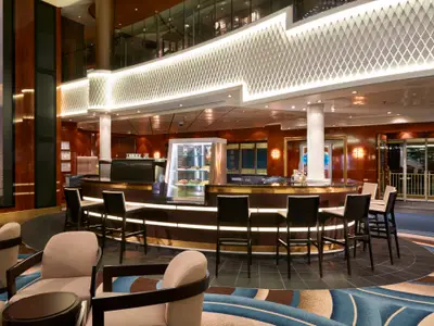 Blick in die moderne Atrium Bar an Bord der Norwegian Gem. Eine runde Bar mit Hockern, eine Kuchentheke und Sitzbereiche prägen den eleganten Raum.