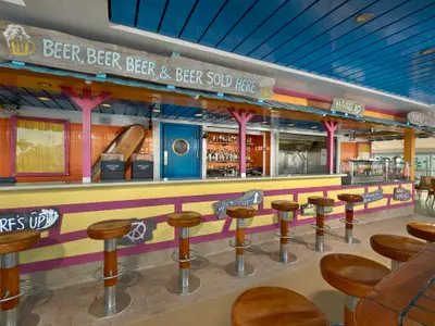 Die Bali Hai Bar an Bord der Norwegian Gem. Ein bunter Tresen mit Barhockern, Surf-Deko und Schilder mit der Aufschrift 'BEER & BEER SOLD HERE'. Im Hintergrund Flaschenregale und eine blaue Tür.
