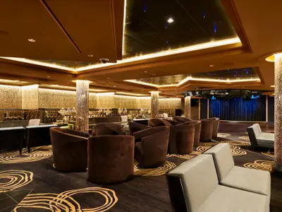 Blick in die elegante Bar-Lounge Gem Bliss an Bord der Norwegian Gem. Braune Sessel und Sofas stehen auf gemustertem Teppich vor einer funkelnden Wand und Bühne.