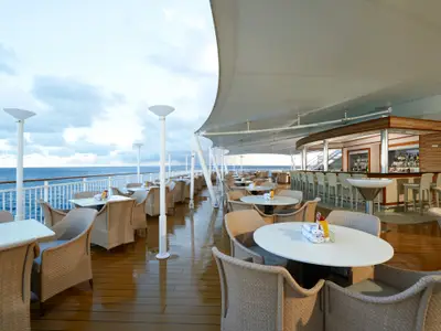 Blick auf das Great Outdoors Bar- und Restaurant auf der Norwegian Gem mit Rattansesseln und Tischen unter einem Sonnensegel, direkt am Meer. Eine lange Bar mit Hockern ist rechts sichtbar.