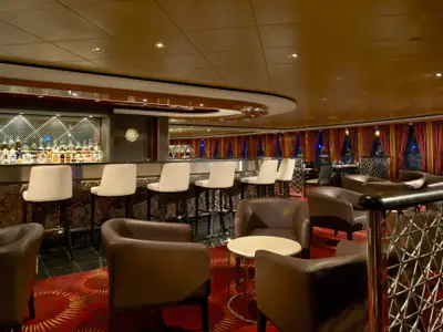 Innenansicht der eleganten Sugarcane Bar-Lounge an Bord der Norwegian Gem. Man sieht eine lange Bar mit zahlreichen Spirituosenflaschen, davor weiße Barhocker und bequeme braune Ledersessel mit kleinen Tischen auf einem roten Teppich. Im Hintergrund sind Fenster mit roten Vorhängen.