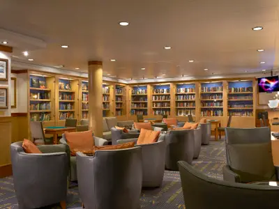 Die gemuetliche Bibliothek an Bord der Norwegian Gem. Holzregale voller Buecher, bequeme Sessel mit orangen Kissen und kleine Tische. Verschiedene Sitzbereiche und Computerarbeitsplaetze laden zum Verweilen ein.