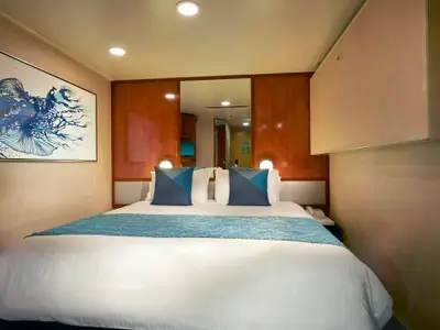 Eine gemütliche Innenkabine an Bord der Norwegian Gem. Im Zentrum steht ein Doppelbett mit weißen Laken, blauer Tagesdecke und zwei Kissen. An der Wand hängt ein großes blaues Kunstwerk mit Fischmotiv.