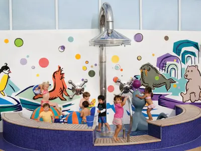 Kinder spielen im farbenfrohen Wasserbereich der Norwegian Gem. Wände mit Arktis-Tieren und Kreisen. Wasserspritzer und Bälle sorgen für interaktiven Spaß in diesem Familienbereich.