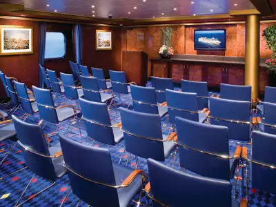 Sitzungsraum an Bord der Norwegian Gem mit Reihen von blauen Stühlen, einem Podium und einem großen Flachbildschirm. Ein Fenster bietet Meerblick.