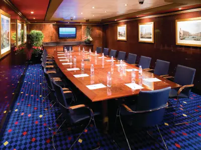 Konferenzraum an Bord der Norwegian Gem mit langem Holztisch, blauen Stühlen, Wasserflaschen und Notizblöcken. Dunkle Holzwände und ein blauer Rauten-Teppich.