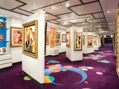Die helle Kunstgalerie an Bord der Norwegian Gem. Zahlreiche Kunstwerke, darunter Gemaelde in Goldrahmen und Skulpturen in Glasvitrinen, sind auf weissen Ausstellungsflaechen zu sehen. Der auffaellige lila Teppich ist mit farbenfrohen Kreisen gemustert und fuehrt durch den Raum.
