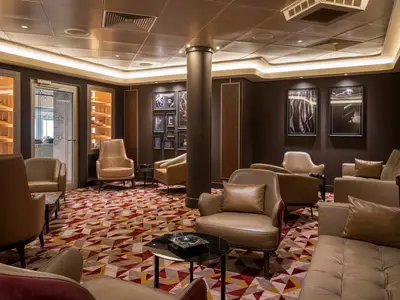 Eine elegante Zigarrenlounge an Bord der Norwegian Gem mit braunen Ledersesseln, Glastischen und einem gemusterten Teppich. Vitrinen praesentieren Zigarren, und die Waende sind in dunklem Braun gehalten.