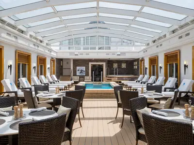 Der luxuriöse, lichtdurchflutete Innenpoolbereich der Norwegian Gem, umgeben von bequemen Liegestühlen und Esstischen mit geflochtenen Stühlen. Im Hintergrund ein Whirlpool und eine Steinfliesenwand sind zu sehen.