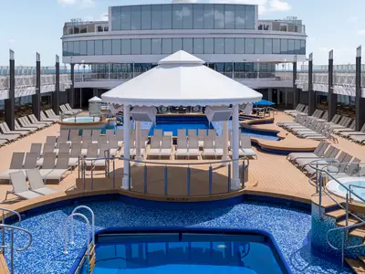Weitblick auf das belebte Pooldeck der Norwegian Gem mit blauen Pools, zahlreichen Liegestuehlen und einer zentralen weissen Buehne. Im Hintergrund sind weitere Decks des Schiffes und das offene Meer erkennbar.