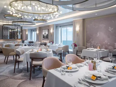 Elegantes Restaurant an Bord der Norwegian Gem. Weiss gedeckte Tische, Stühle und Meerblick durch Fenster. Stilvolle Deckenleuchten und Wandkunst.