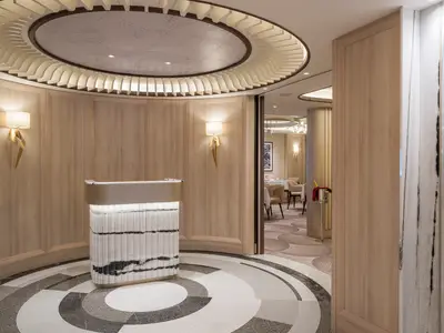 Empfang des Le Bistro an Bord der Norwegian Gem. Beleuchtetes Marmorpult vor Holzwänden. Moderne Wand- und Deckenleuchten. Tische im Hintergrund.