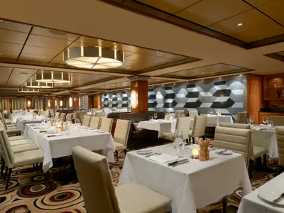 Blick in das elegant gedeckte Spezialitätenrestaurant Cagney's an Bord der Norwegian Gem. Zahlreiche Tische mit weissen Tischdecken, Geschirr und Gläsern sind zu sehen, umgeben von bequemen Stühlen und gepolsterten Bänken. Eine Wand ist mit einem modernen geometrischen Muster verziert, während die Decke aus Holzpaneelen und markanten Leuchten besteht.