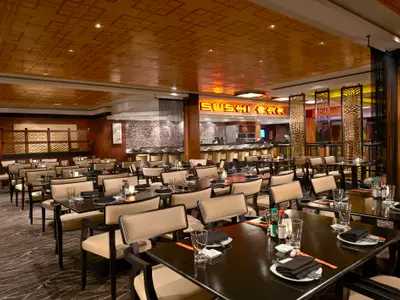 Innenansicht des eleganten Orchid Garden Spezialitätenrestaurants an Bord der Norwegian Gem mit vielen gedeckten Tischen und Sushi Bar. Der Raum ist in warmen Brauntönen gehalten.