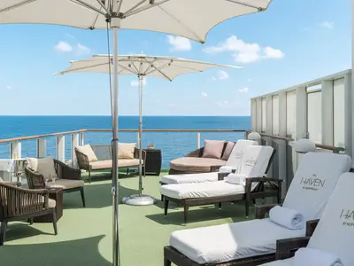Ein luxuriöses, privates Sonnendeck von The Haven an Bord der Norwegian Gem. Es zeigt bequeme Loungemöbel, Sonnenschirme und einen weiten Blick auf das offene Meer unter blauem Himmel mit einigen Wolken. Eine Champagnerflasche und Gläser stehen bereit.