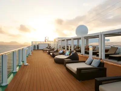 Das Sonnendeck der Norwegian Gem bei Sonnenuntergang, mit eleganten Lounge-Möbeln und Meerblick. Im Hintergrund sind Berge und ein leuchtender Himmel zu sehen.
