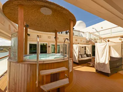 Ein luxuriöses Sonnendeck der Norwegian Gem mit großem Whirlpool unter einem Reetdach, eleganten Lounge-Cabanas und Meerblick auf eine Küstenlandschaft.