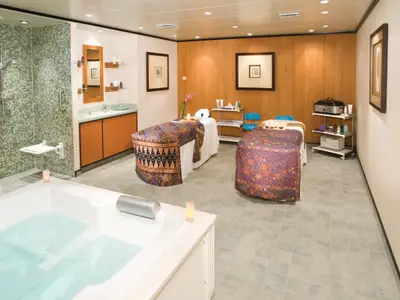 Ein luxuriöser Spa-Behandlungsraum an Bord der Norwegian Gem mit zwei Massagebetten, einem Whirlpool, einer begehbaren Dusche mit Mosaikfliesen und Holzelementen.