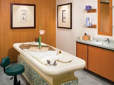 Spa-Behandlungsraum an Bord der Norwegian Gem. Zeigt Massageliege, Waschbecken mit Holzschränken, Pflegeprodukten, asiatische Kunst und Blume. Entspannte Atmosphäre.