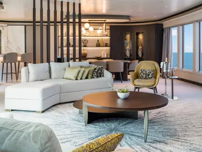 Luxuriöse Suite der Norwegian Gem mit Meerblick. Ein Wohnbereich mit beiger Eckcouch, Sessel, Couchtisch und eine Bar sind zu sehen. Die Einrichtung ist modern und elegant.