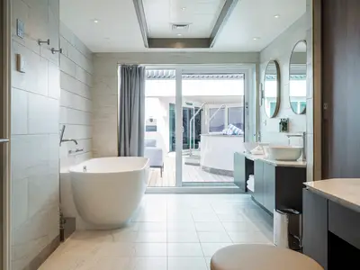 Modernes Bad in einer Suite der Norwegian Gem mit Badewanne und Doppelwaschbecken. Eine Glastür führt zu einem privaten Balkon mit Whirlpool und Loungesesseln.