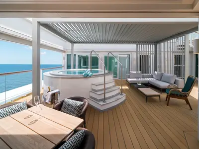 Ein luxuriöser Außenbereich einer Suite an Bord der Norwegian Gem mit privatem Whirlpool, gemütlichen Sitzmöbeln und weitem Blick auf das Meer unter blauem Himmel.