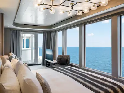 Luxuriöse Suite der Norwegian Gem mit großem Bett im Vordergrund. Bodentiefe Fenster bieten weiten Meerblick. Sitzecke mit Sessel, TV und Zugang zum Balkon links. Modernes Design.