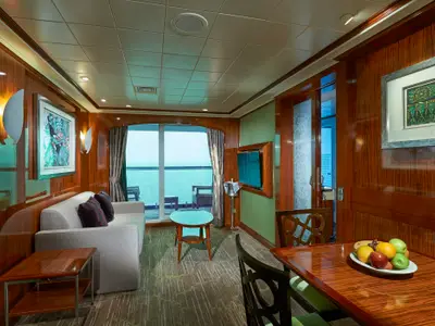 Blick in eine geräumige Suite an Bord der Norwegian Gem mit elegantem Holzinterieur, Sofa, Esstisch und großen Fenstern zum Meer hinaus. Ein Obstkorb steht bereit.